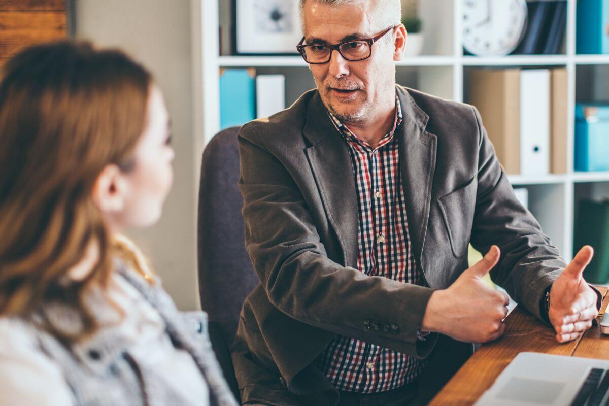 The picture is a man in a suit explaining and a girl listening to him with a laptop on the table referring to seeking a financial mentor for accountability that can help you stay on track and offer advice to rebuild your money habits.