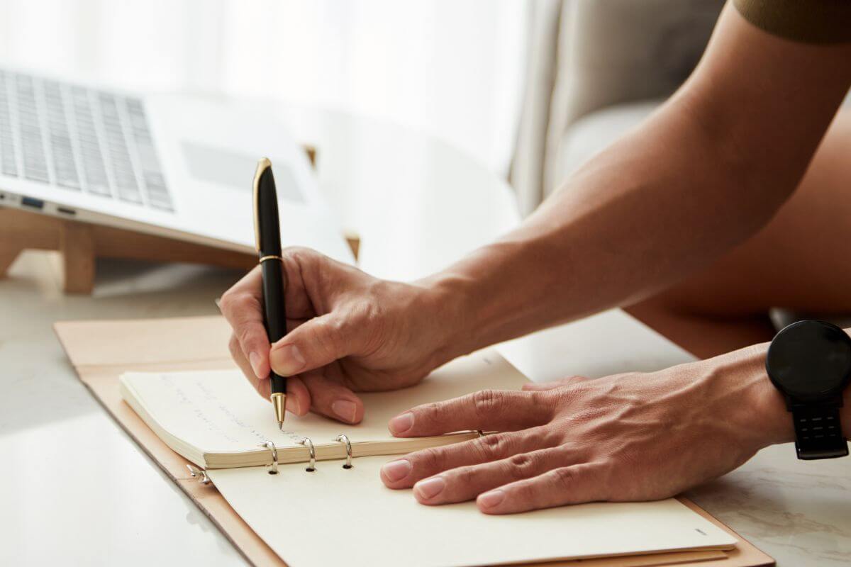 The picture tells about a laptop and a man's hand and writing on the notebook indicating that practicing daily gratitude journaling helps shift your mindset and reminds you there's more to life than financial stress.