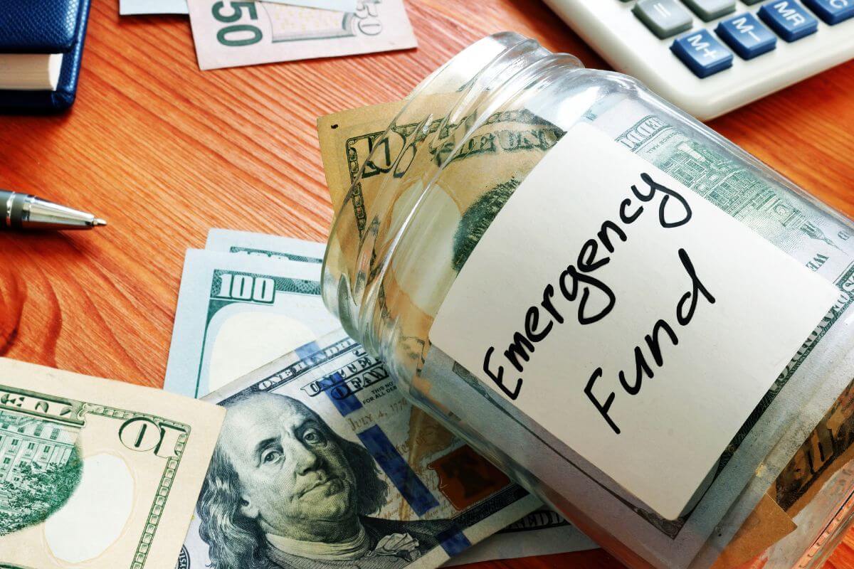 The photo shows dollar bills, a pen, a calculator, and a jar filled with dollar bills and with noted indicating to build an emergency fund will give you peace of mind and educe stress when unexpectred costs ccame up.