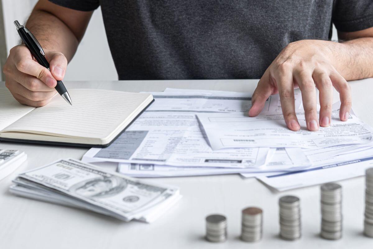 The image of stock coins, dollar bills a calculator, papers, and a man's hand and writing on the notebook while pointing the papers indicating to cut discretionary expenses ruthlessly that every dollar saved toward fixing your financial situation.