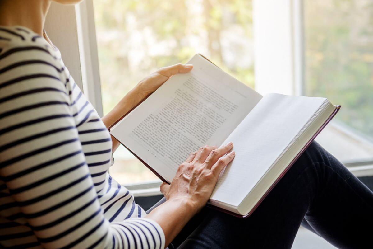 The picture of a girl reading a book at the window view refers to reading books or articles on personal finance to build the knowledge that can help you avoid future setbacks and take control of your financial health.