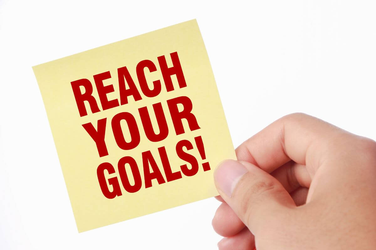 The photo shows a man's hand holding a piece of paper with notes indicating that knowing how you want to reach your goals gives you direction and makes reaching your goals feel manageable.
