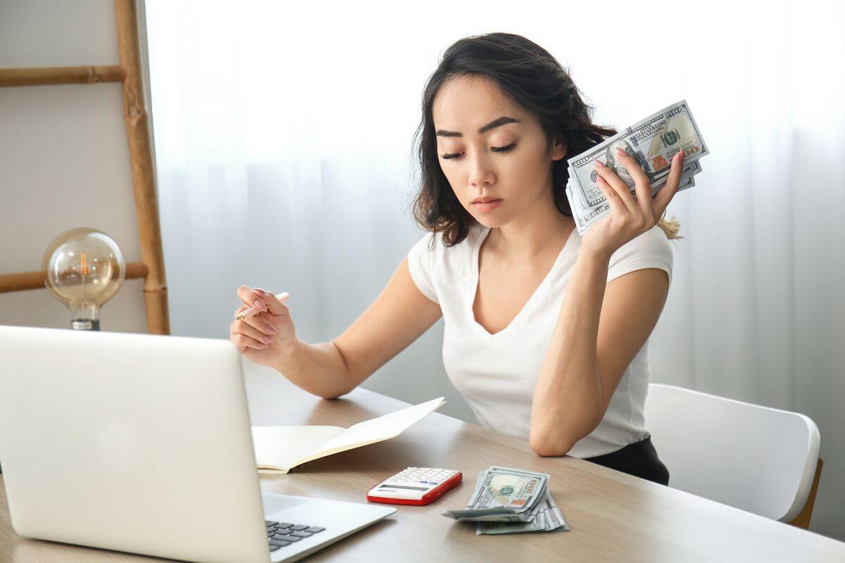 The photo of a laptop, a calculator, dollar bills, a notebook, and a girl holding dollar bills and a pen means to make your money work for you by putting your money into accounts or investments that grow it over time to build wealth.