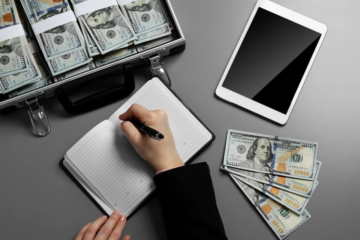 The image shows a tablet, dollar bills, a container with bucks of dollar bills, and a girl's hand and writing on the notebook which means learning how to become independently wealthy helps you take control of your finances and work toward independence.
