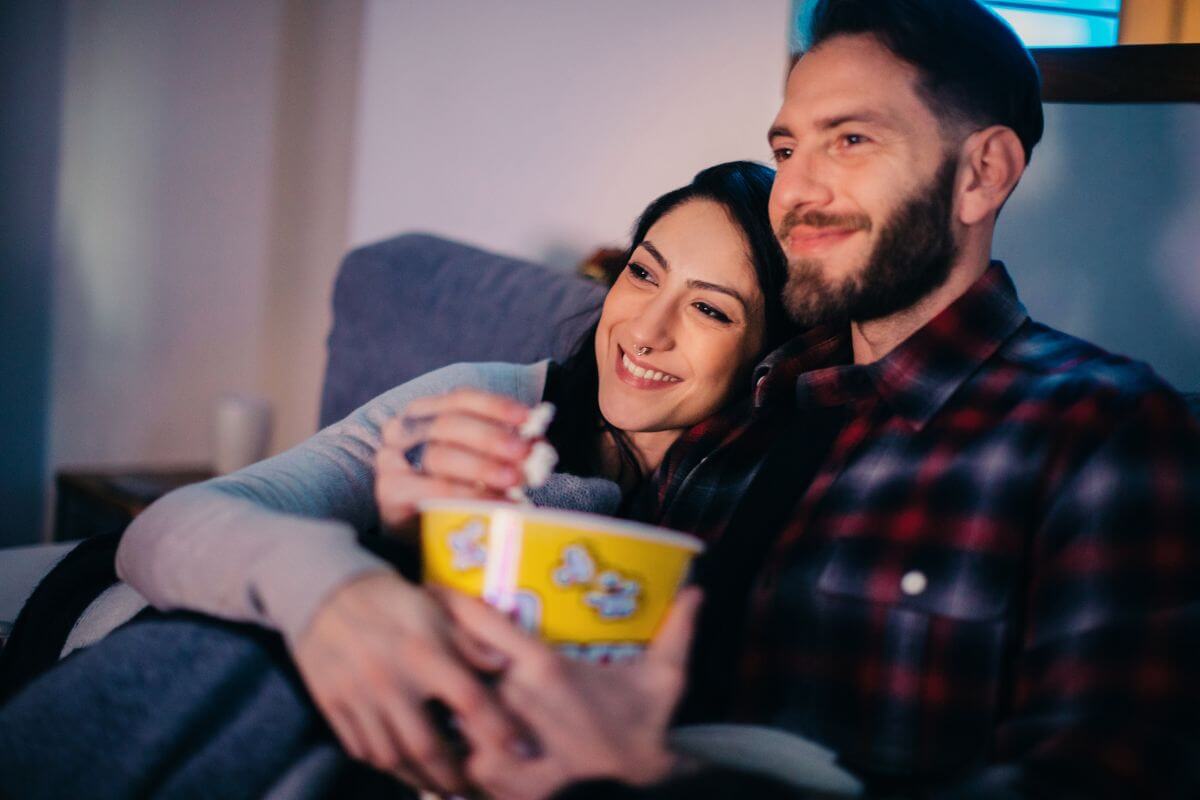 The picture shows a couple with a popcorn indicating to to plan a movie night at home which an easy way to enjoy time together while skipping the cost of going to a theater.