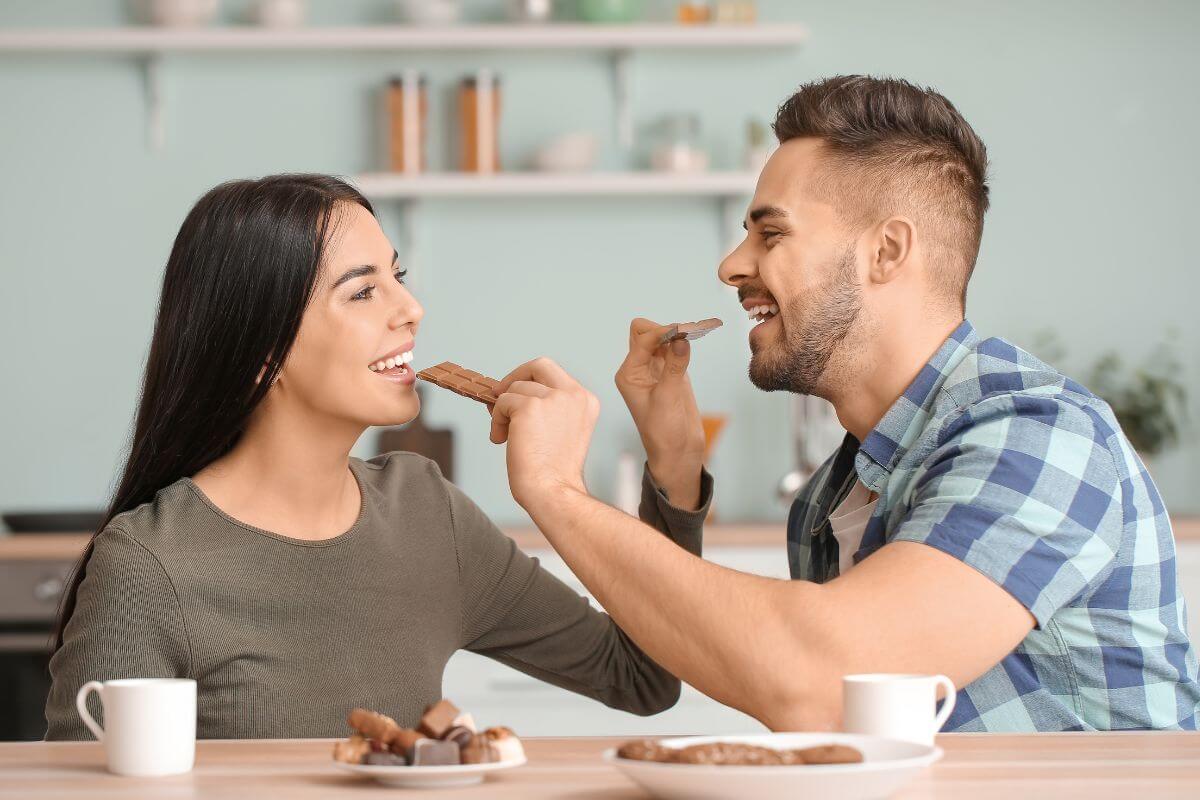 The image shows chocolates on the plate, mugs, and a couple eating chocolates which refers to a DIY chocolate tasting experience at home, a fun way to treat yourselves without buying pricey desserts.