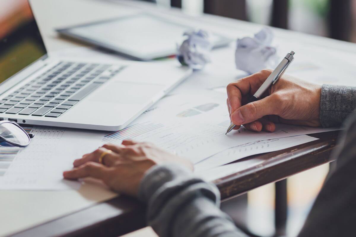 The image shows of a laptop, crumpled papers, and a girl's hand writing on the papers which means to write down all your income sources helps you see what you're working with so you don't spend more than you make.