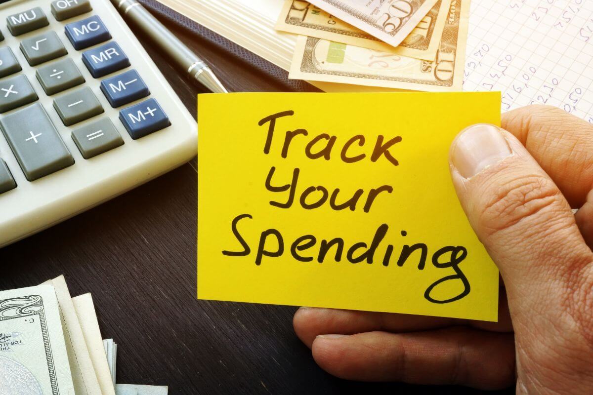 The image shows a calculator, a pen, dollar bills, a notebook, and a man's hand holding a piece of paper with notes which means tracking all your spending daily helps you stay in control and avoid surprise expenses.