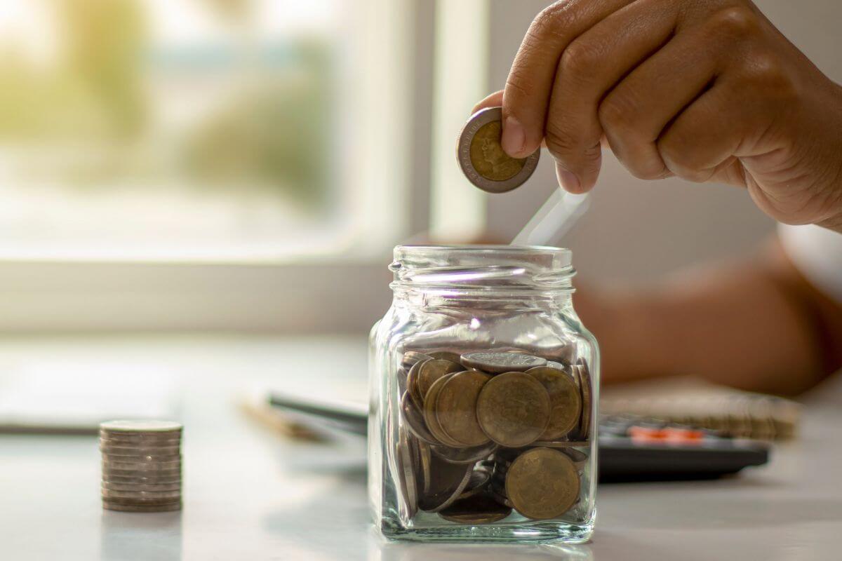 The image shows coin stock,  a calculator and a man's hand dropping a coin into the jar filled with coins indicating that setting aside a small amount for unexpected expenses helps you handle surprises without stress.