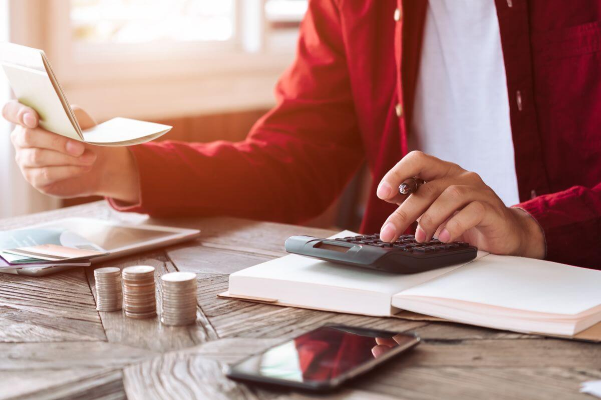 The photo shows coin stocks, a phone, a tablet device, a notebook, and a man calculating while holding a bank book which means sticking to a simple budget method helps balance needs, wants, and savings without overcomplicating things.