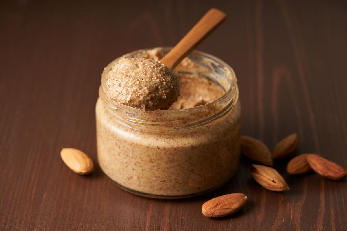 The image shows almonds on the table and a jar with a wooden spoon referring to fancy nut butter costs much more than peanut butter, buying plain nut butter or making your own can save a lot.