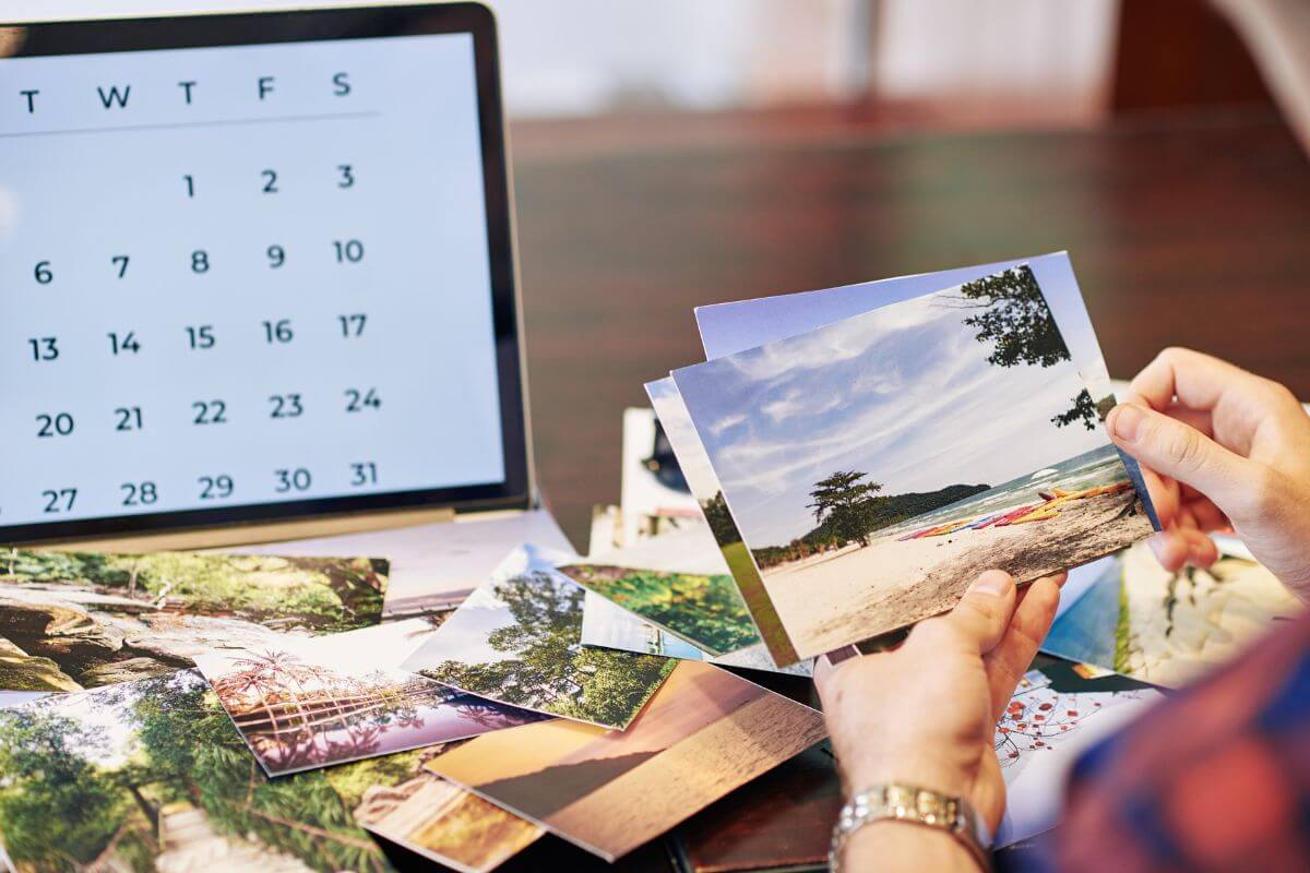 The picture tells about a laptop with a calendar and a girl's hand picking up photos referring to a DIY photo calendar that makes every month special with memories of you both. 