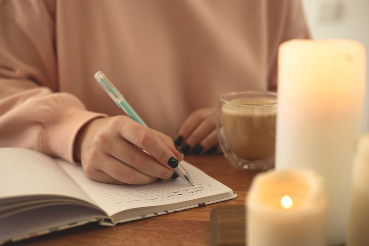 The image shows candles, a coffee, and a girl writing on the notebook which means a journal with prompts for them to fill out makes a meaningful and personal gift.