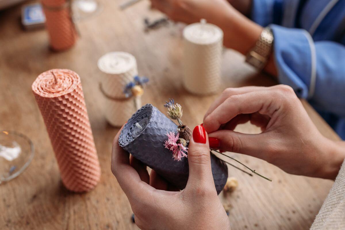 The photo shows a girl's hand designing the candle with flowers and candles on the table indicating that making a DIY candle with their favorite scent is a warm and thoughtful gift.