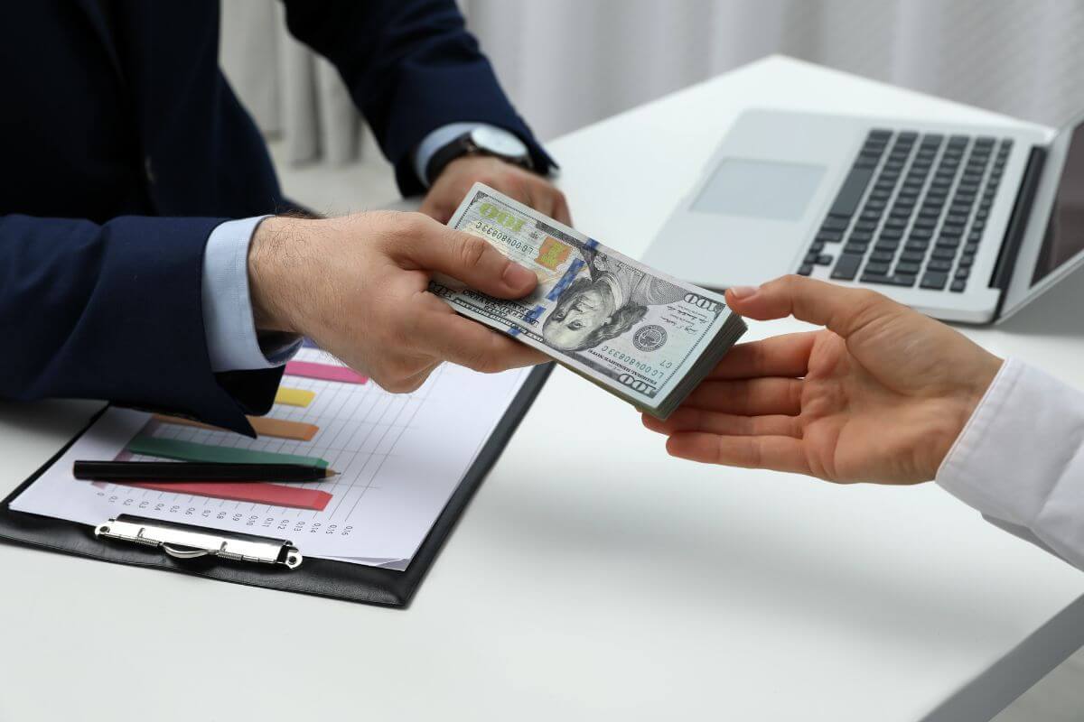 The photo shows a laptop, a clipboard with a graph, a pen, and a man in a suit handing the dollar bills to the man's hand which means making large financial decisions without consulting you shows a lack of respect and teamwork, healthy relationships involve financial transparency and share decision- making.
