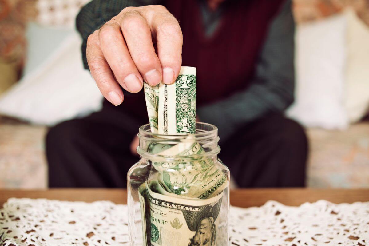 The photo shows a senior's hand dropping dollar bills inside a jar filled with dollar bills which means early retirement depends on their savings, expenses, and retirement goals to become financially ready.