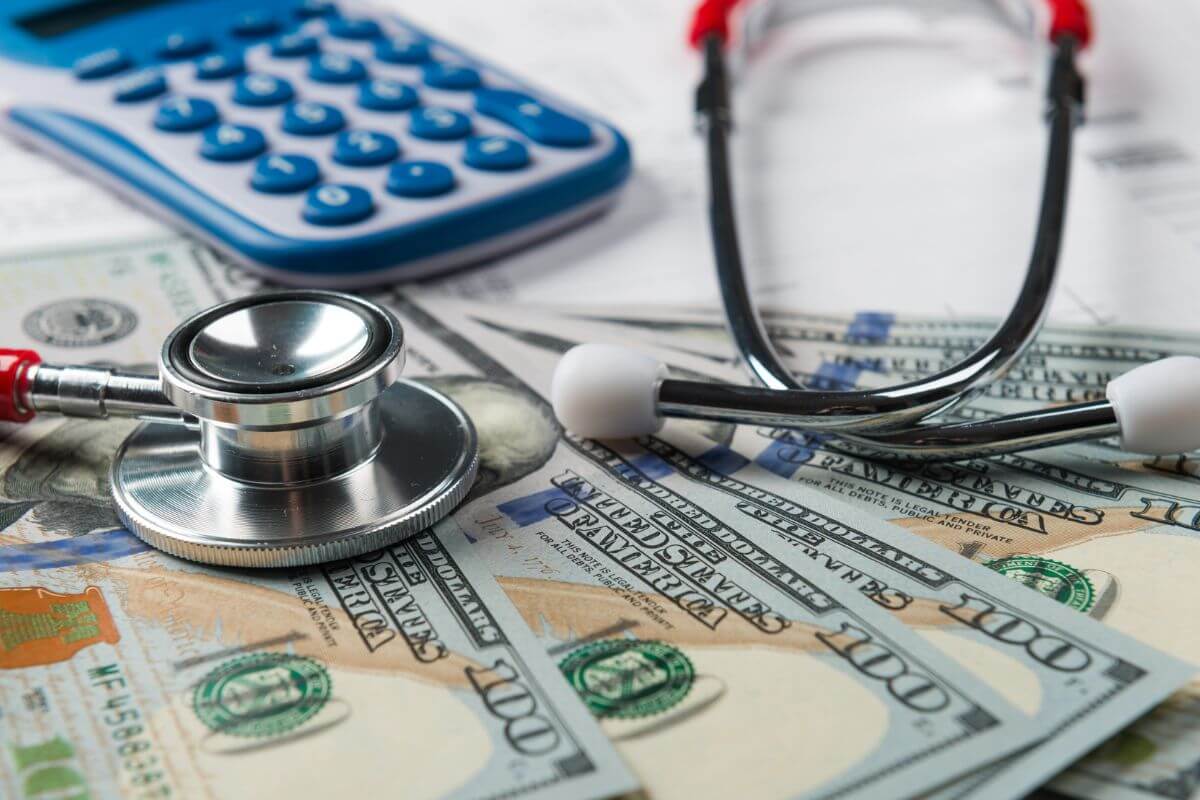 The photo shows a calculator, a stethoscope, and dollar bills referring to healthcare costs that should be factored in to avoid financial difficulties in the future.