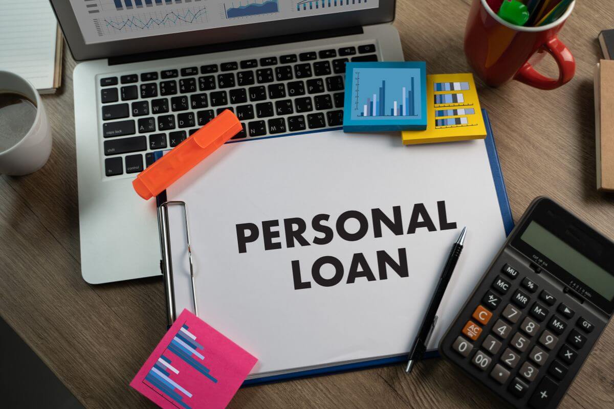 The picture shows a laptop, coffee, pen, and sticker notes with graphs, a calculator and a clipboard with notes referring to personal loans may have lower interest than credit cards, but they still take a chunk out of your monthly income.