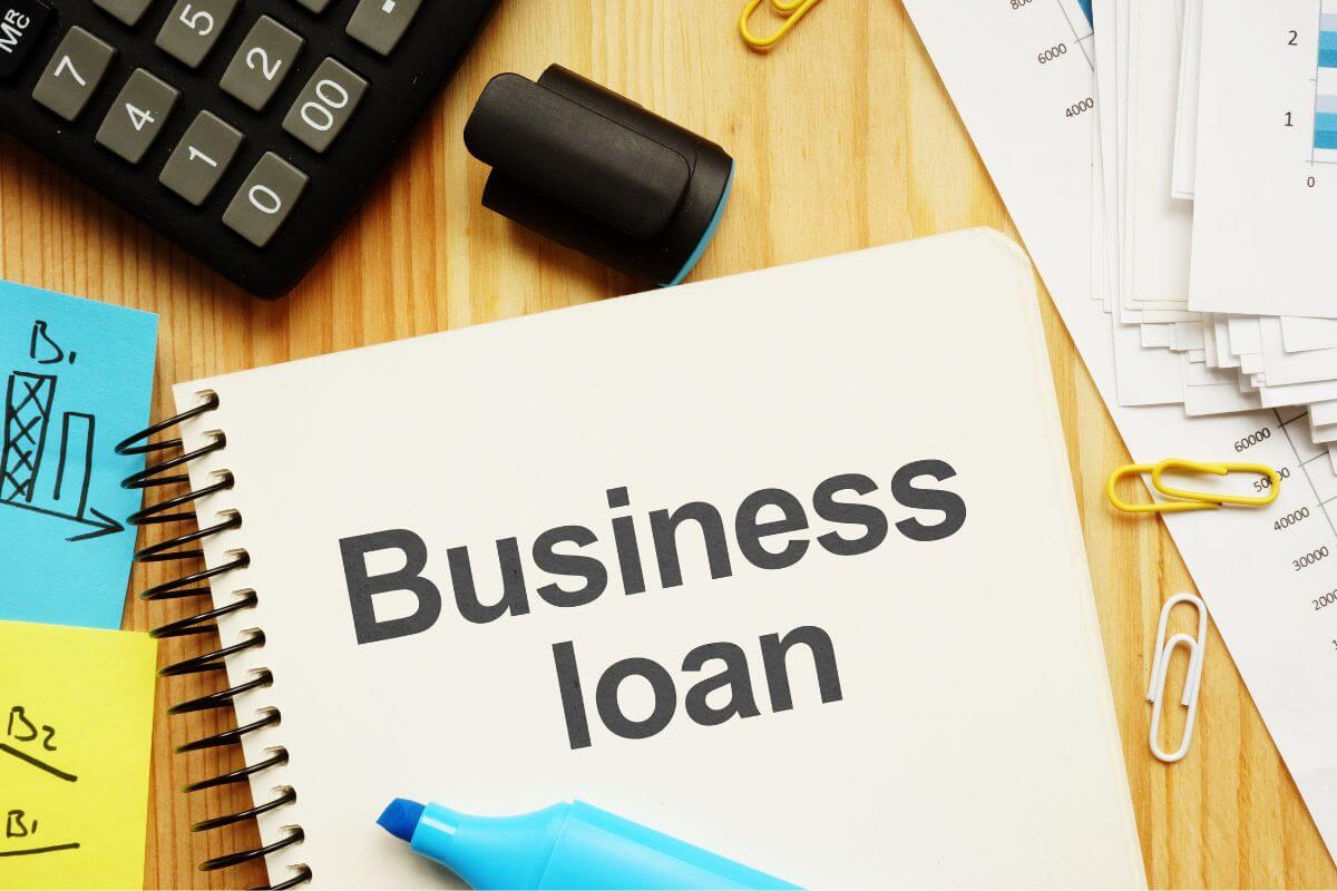 The image shows a calculator, papers, sticker notes, a marker, clips, and a notebook with notes referring to business loans that could follow you into retirement, paying it off before leaving the workforce protects your personal savings.