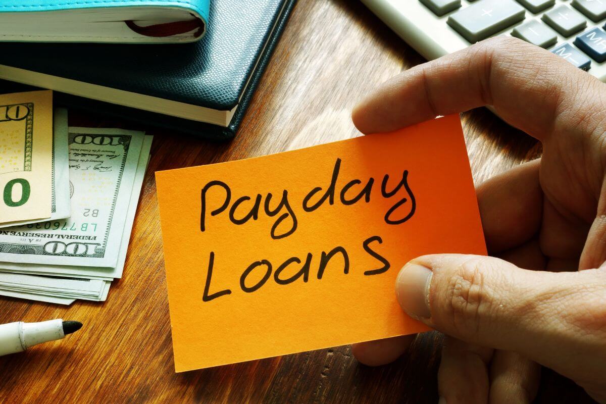 The photo tells about dollar bills, notebooks, a calculator, a marker, and a man's hand holding a piece of paper with notes referring to payday loans come with sky-high interest rates that can trap you in a cycle of debt, paying them off quickly saves you from long-term financial trouble.