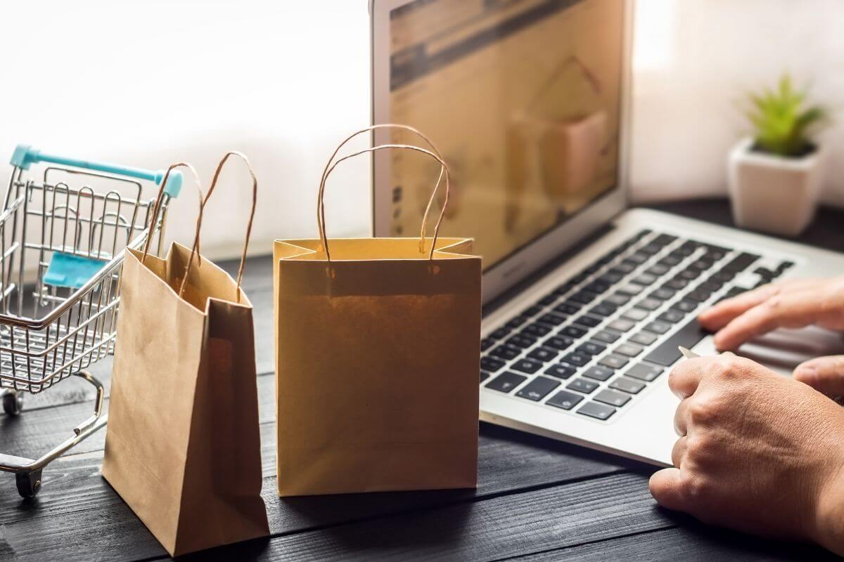 The image shows a mini cart, shopping bags, and a girl's hand using a laptop which means the pledge to buy less helps break the habit of buying things just and making better financial stories.