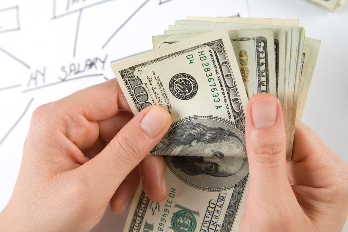 The image shows a girl's hand counting dollar bills and a piece of paper with notes indicating to live on last year's salary to grow savings fast and put the extra money straight into savings or retirement.