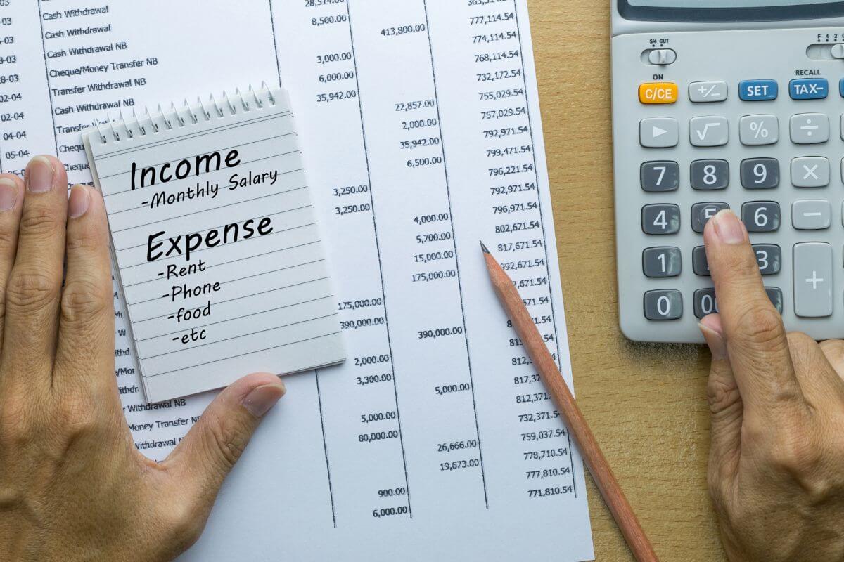 The photo tells about a piece of paper with notes, a pen, a mini notebook with notes, and a man's hand using a calculator indicating to start small by tracking your income and expenses to understand where your money goes.