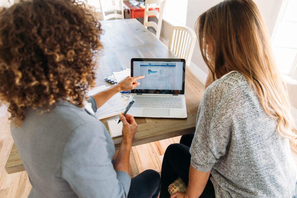The image shows a girl in the suit explaining the content on the laptop to a girl referring to consider reaching out to a financial advisor to explain things in a way that makes sense.