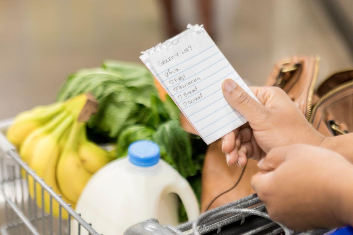 The picture shows a cart filled with fruits, vegetables, a gallon of milk, a bag, and a girl's hand holding a paper with notes indicating to start your budget grocery list to save money and avoid food waste.