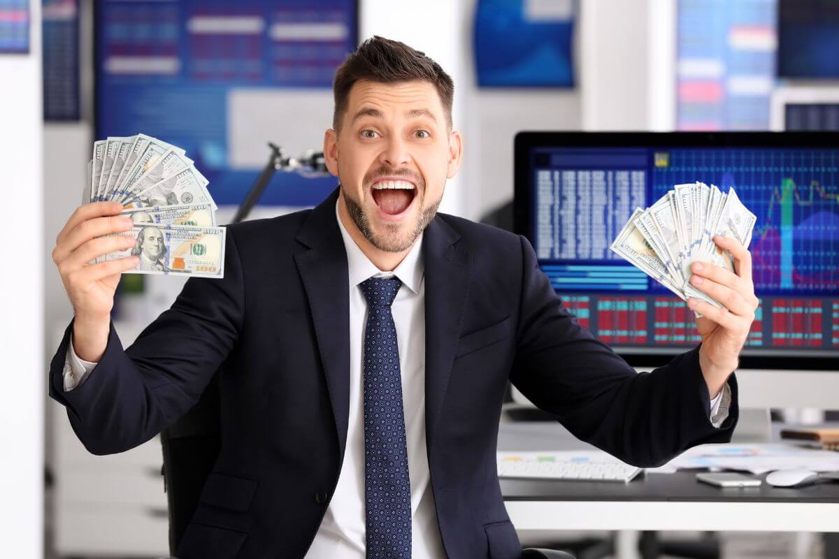 The image shows a computer with a screen display and a man's hand holding dollar bills referring to a stock trader can be a great way to make money without talking to people all day.