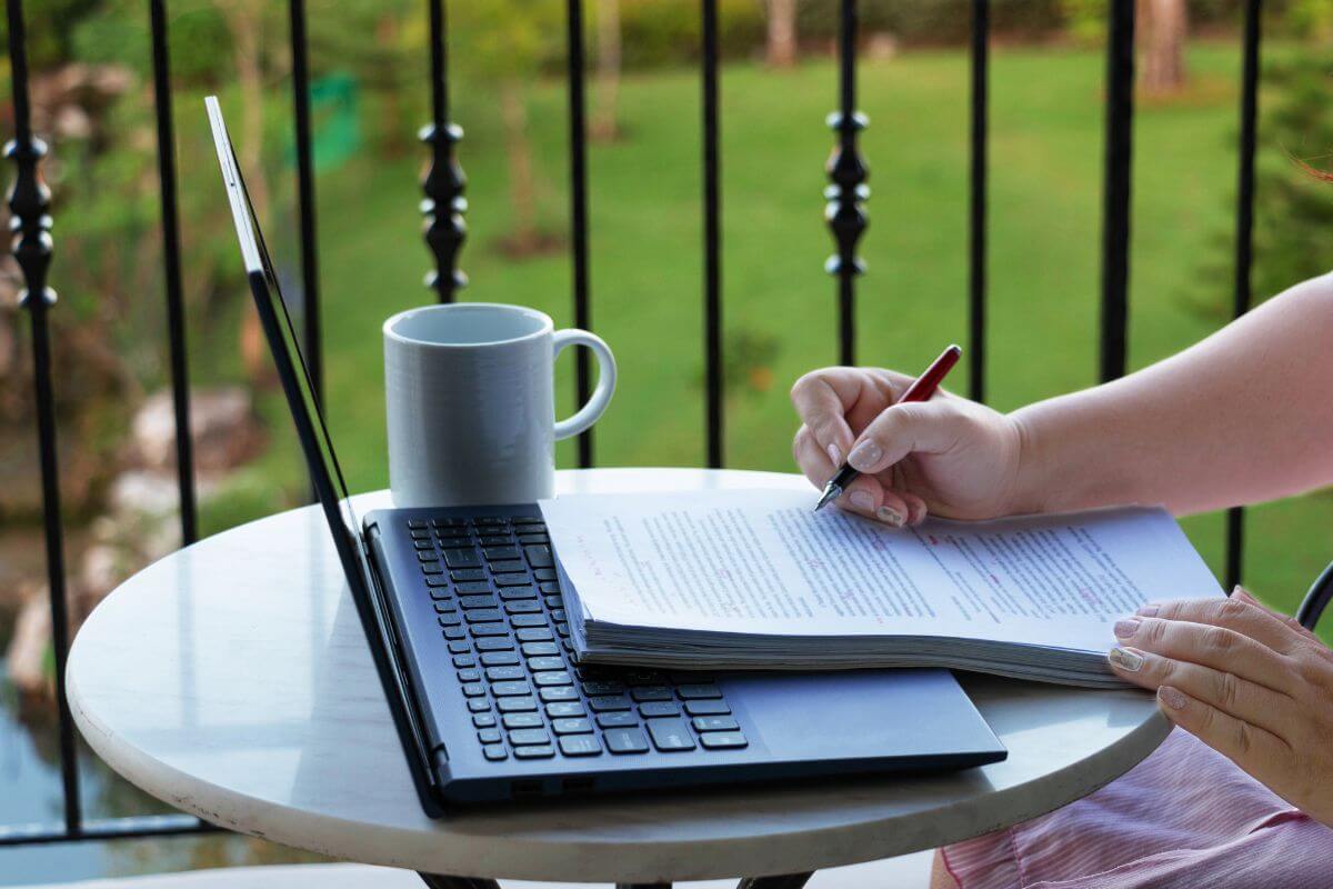 The image shows a mug, a laptop, and a girl reading the papers while holding a pen which means proofreading and editing can be a quiet way to make money online.