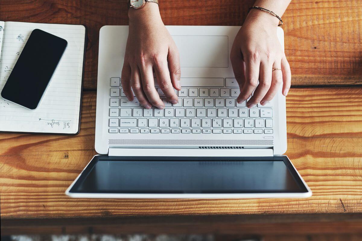 The photo of a phone, a notebook, and a girl's hand using a laptop indicates that a transcription can earn money, a perfect job for those detail-oriented and enjoy working quietly.