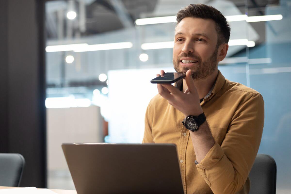 The image shows a laptop and a man recording his voice through a phone which means voiceover work for commercials and more can be a great job from home on your own schedule.