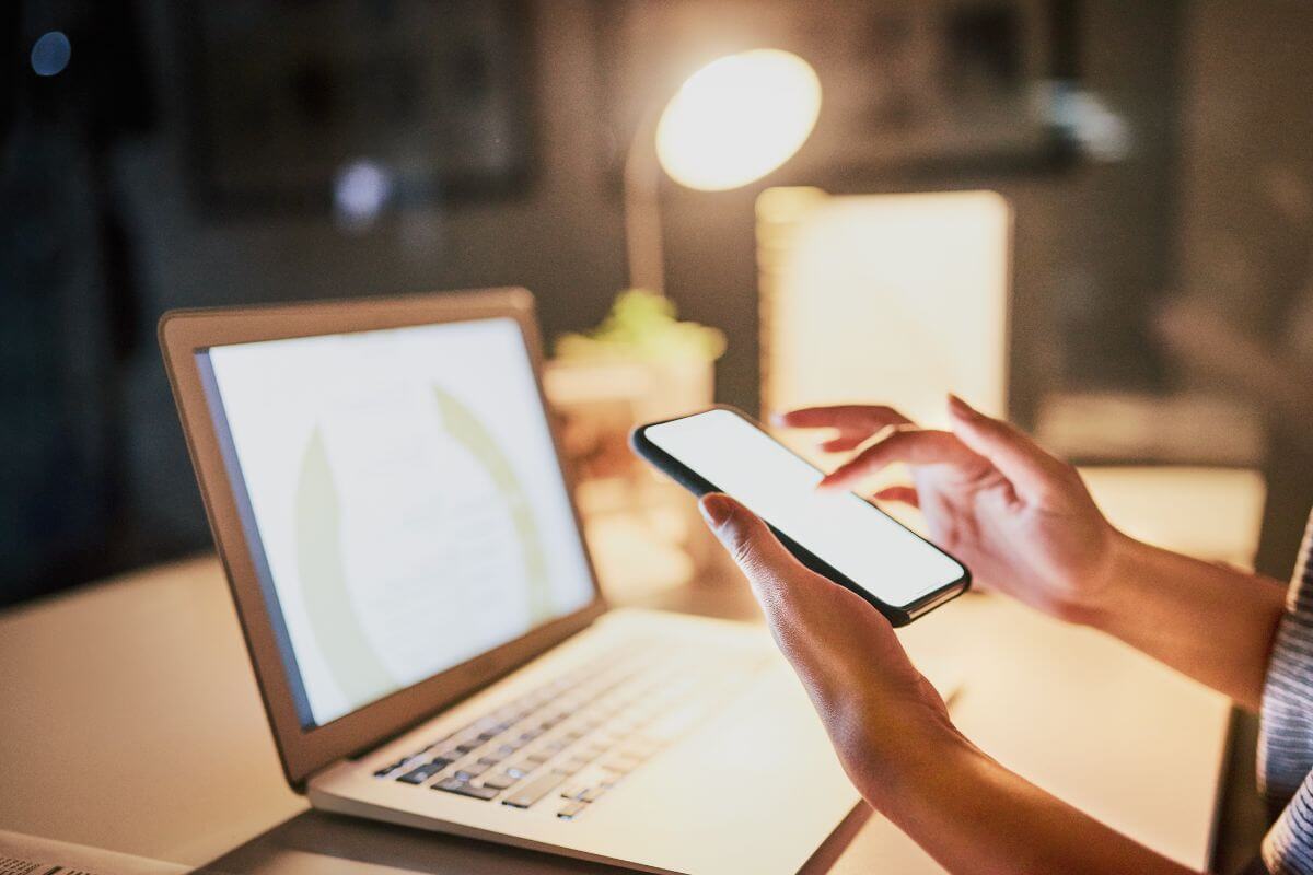 The photo tells about a laptop and a girl's hand using a phone referring to social media management can be a great work-from-home job, working independently with minimal interactions.