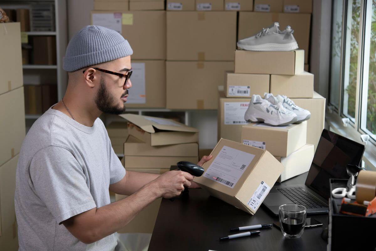 The image shows boxes with shoes, pens on the table, a coffee, a laptop, and a man scanning the code indicating that an E-commerce seller lets you sell without direct customer interaction.