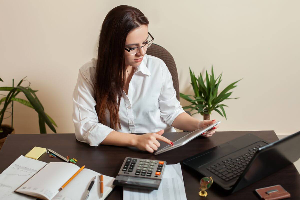 The image shows papers on the table, notebooks, a laptop, pens, a calculator, a laptop, and a girl using a tablet device which means bookkeeping perfect for detail-oriented that can earn while from home.