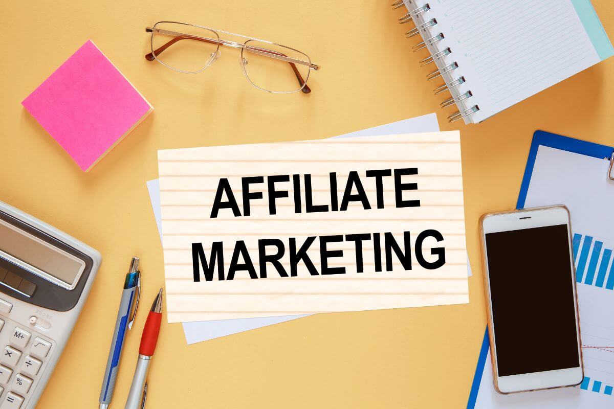 The image shows a calculator, sticker notes, an eyeglass, a phone, a clipboard, and papers with notes referring to affiliate marketing is a great passive income option that can make money even at home.