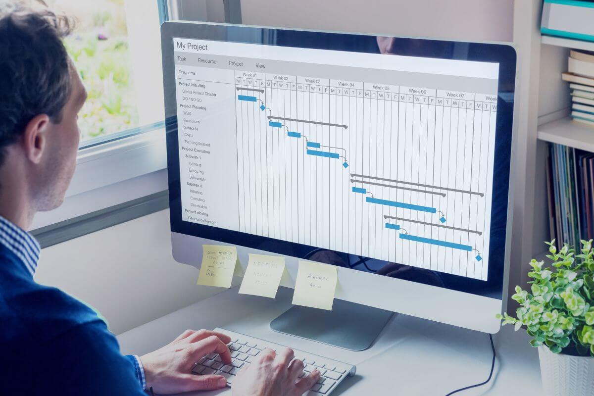 The photo shows a man using a computer with a screen display which means that remote project management can be a good earning at home by keeping projects organized for a team.