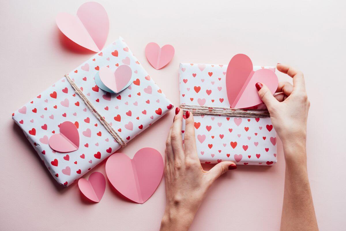The picture shows a girl's hand designing the gift boxes with hearts shaped papers referring to doing a DIY gift or create something personal is an easy way to make Valentine's Day special without spending.