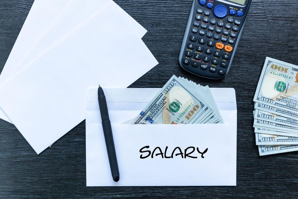 The picture shows envelopes, dollar bills, a calculator, a pen, and an envelope with dollar bills and a note which means by 40, target 3x your salary in savings by cutting unnecessary spending, boost savings, and invest wisely. 