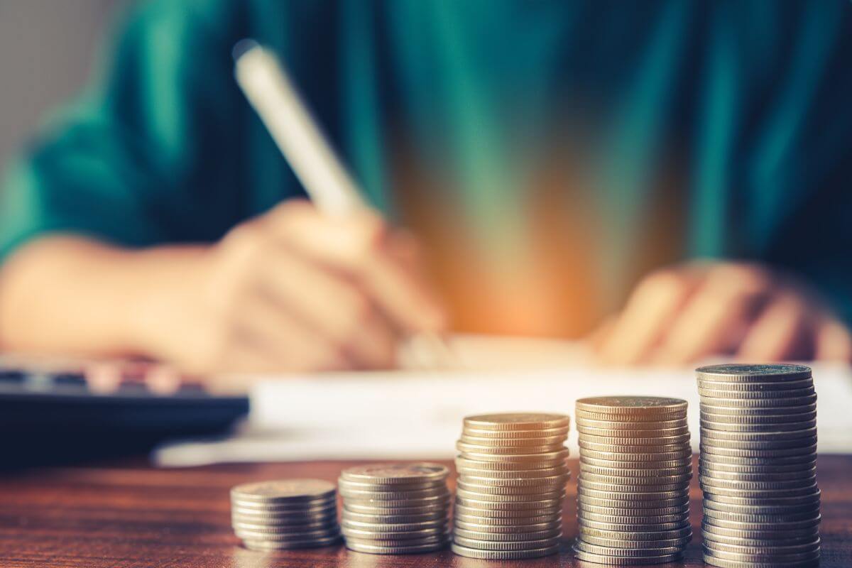 The image shows coin stocks, a calculator, and a girl writing on the notebook which refers to flipping the budgeting rule by flipping to 20/30/25 which can supercharge retirement goals.