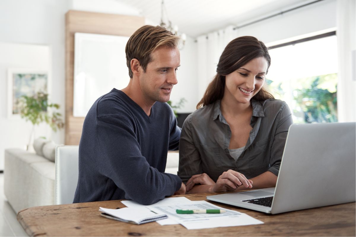 The photo tells about papers, a notebook, a marker, and a couple using a laptop, indicating the time to reach those goals to reach financial freedom and build a bright future together. 