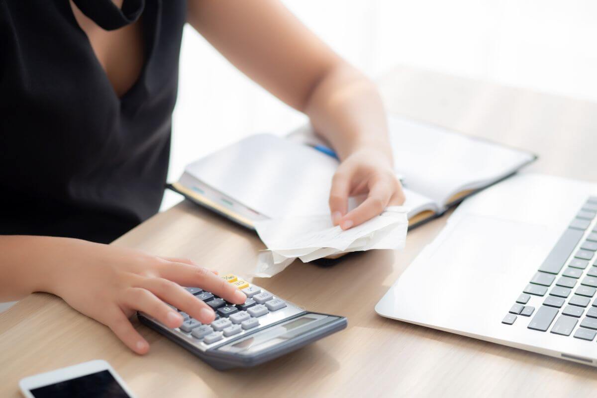 The picture shows a laptop, a phone, a notebook, and a girl holding papers while calculating which means cut-out phantom expenses can quietly drain your bank account.