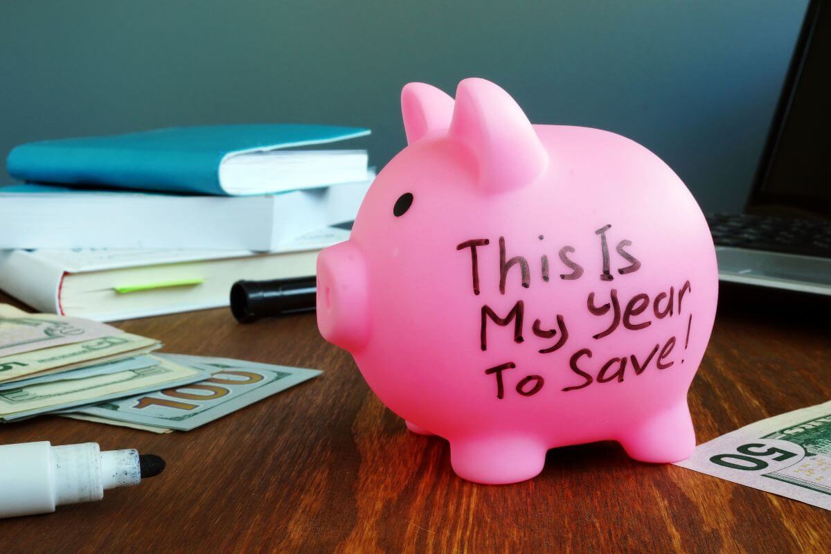 The picture shows notebooks, dollar bills, a marker, a laptop, and a piggy bank with notes which means try a savings challenge or plan a no-spend challenge to help you grow your account without feeling overwhelmed.