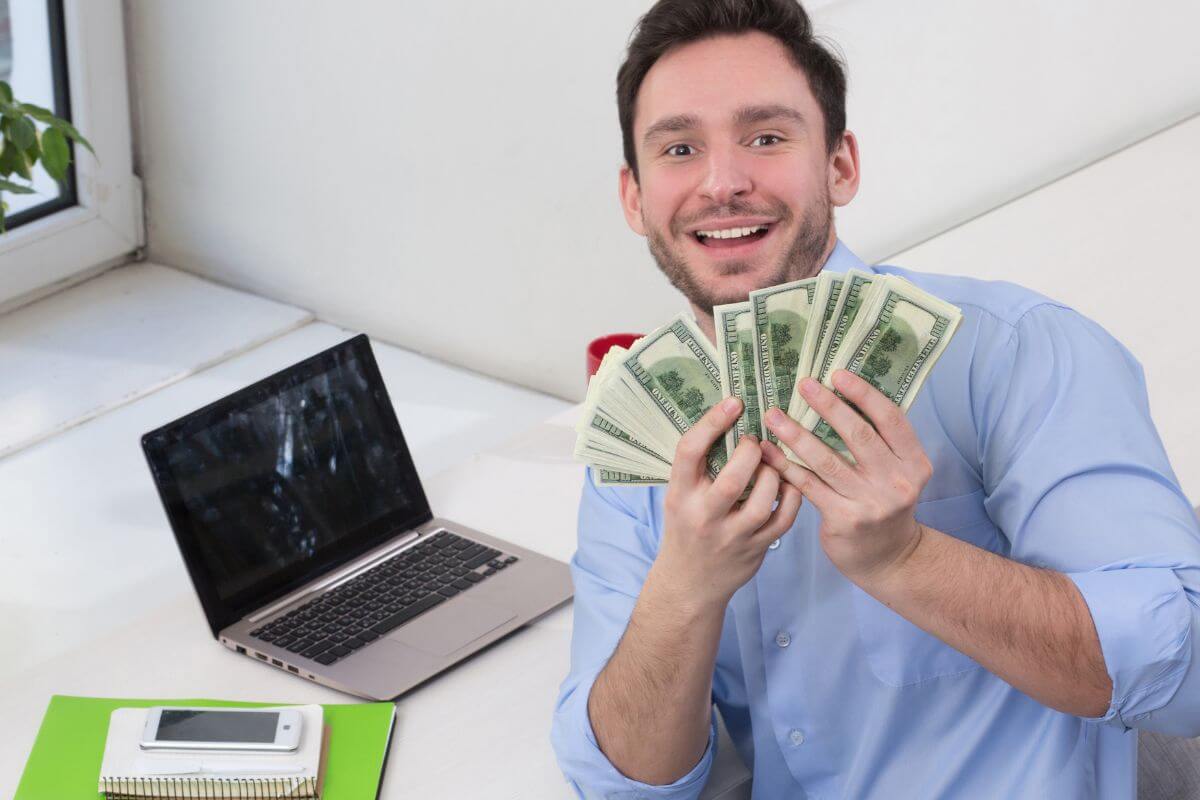The photo shows a laptop, a phone, a notebook, and a man holding dollar bills which means turning a hobby into extra income that can bring in extra cash and a smart way to grow financial security while doing something you love.