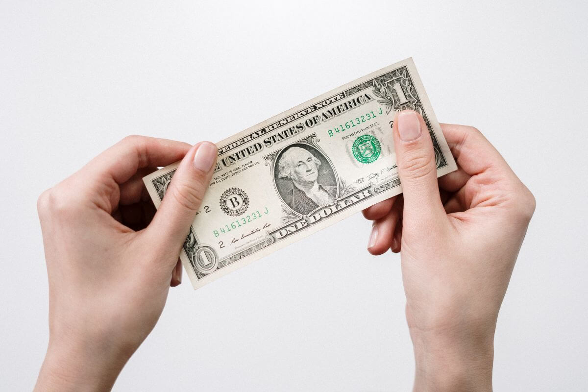 The photo tells about a girl's hand holding a dollar bill which means challenging yourself to save at least 1$ more each day can turn into big money for retirement savings.