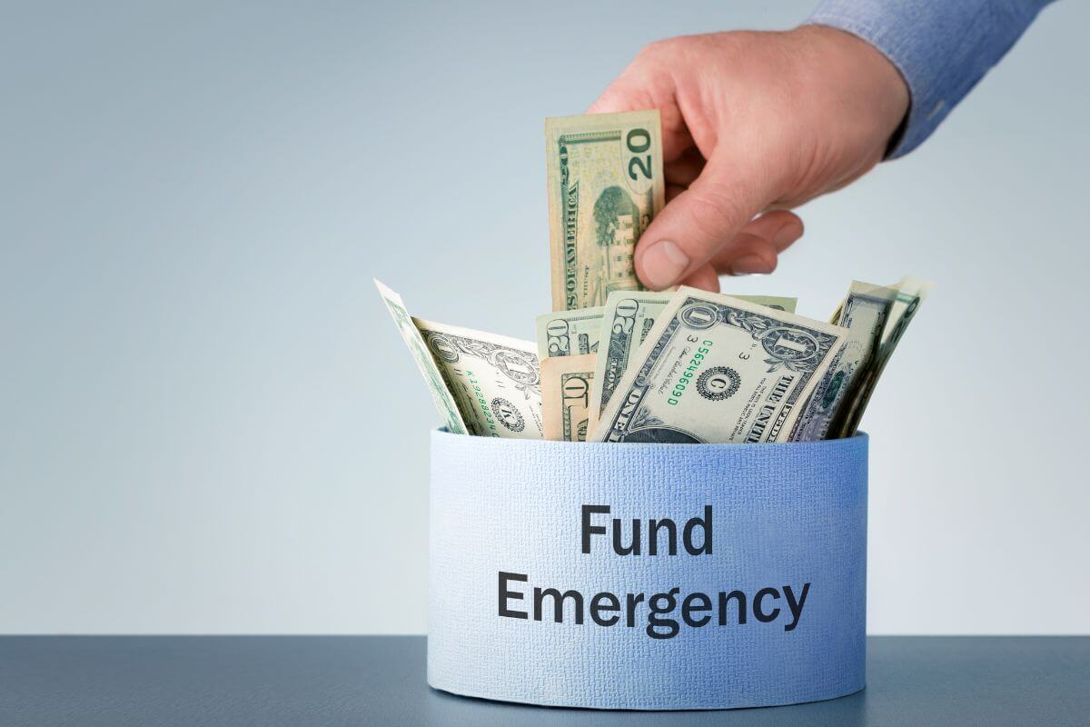 The image shows a container with dollar bills and notes and a man's hand putting a 20 dollar bill to it which means building an emergency fund with 6-12 months of expenses to avoid financial stress on unexpected costs.