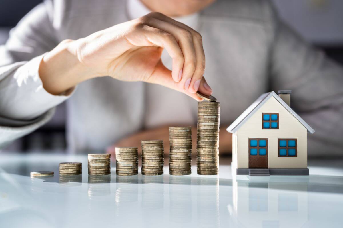 The image shows coin stocks, a mini house, and a man's hand putting a coin referring to saving for a home or real estate investment to grow your money.