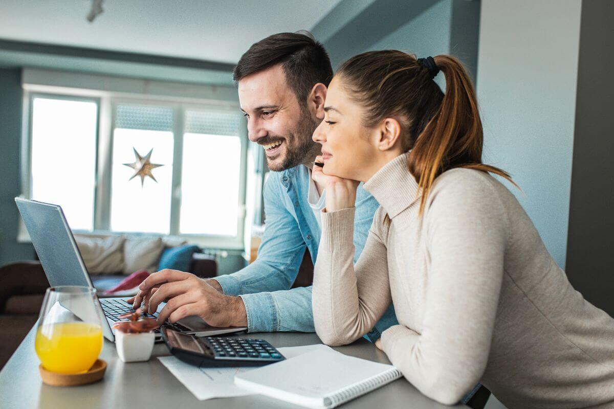 The photo shows a notebook, paper, a calculator, juice, and a couple using a laptop indicating to set a joint monthly budget and track the expenses together to avoid overspending.