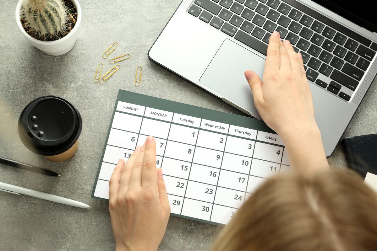 The photo shows a plant, pins, a coffee, pens, and a girl using a laptop while pointing at a calendar, referring to setting a timeline for achieving financial independence to stay motivated and achieve goals more easily.